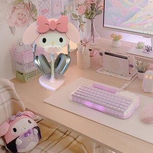 May include: A pink Hello Kitty themed headphone stand with a white base and silver headphones. The stand is on a light pink desk with a white keyboard and a white mouse.