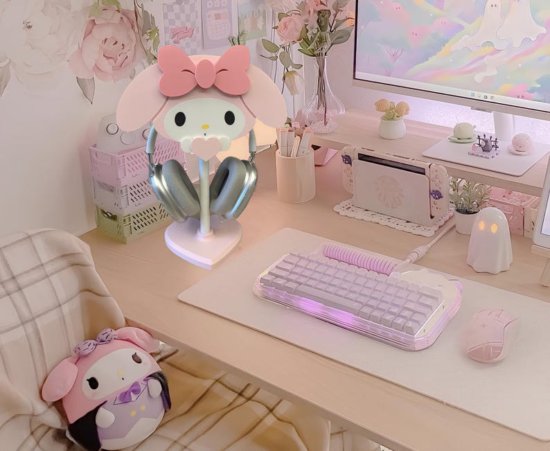 May include: A pink Hello Kitty themed headphone stand with a white base and silver headphones. The stand is on a light pink desk with a white keyboard and a white mouse.