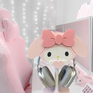 May include: Pink Hello Kitty themed headphone stand with a heart-shaped base. The stand is made of wood and features a cute Hello Kitty face with a pink bow.