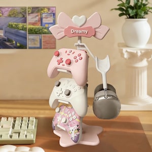 May include: A pink and white gaming controller stand with three controllers and a headset. The stand has a bow and heart design with the word "Dreamy". The controllers are pink, white, and a patterned pink and purple.
