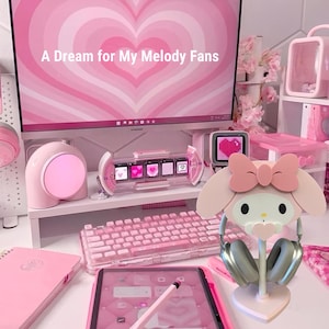 May include: A pink and white My Melody headphone stand with a pink bow. The stand is on a white desk with a pink keyboard, a pink notebook, and a pink computer monitor with the text "A Dream for My Melody Fans".
