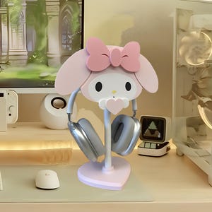 May include: Pink Hello Kitty themed headphone stand with a heart-shaped base. The stand has a white post and two grey headphones are hanging on it.