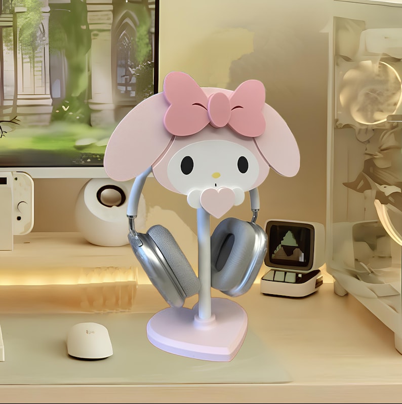 May include: Pink Hello Kitty themed headphone stand with a heart-shaped base. The stand has a white post and two grey headphones are hanging on it.