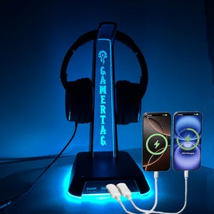 以下が含まれることがあります： 青いLEDライトが付いた黒いヘッドセットスタンド。スタンドには2つの充電ポートがあり、2つのスマートフォンを充電しています。スタンドには「GAMERTAG」と印刷されています。