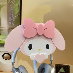 May include: Pink and white Hello Kitty headphone stand with a pink bow. The stand has a heart-shaped cutout in the front and two hooks for headphones.