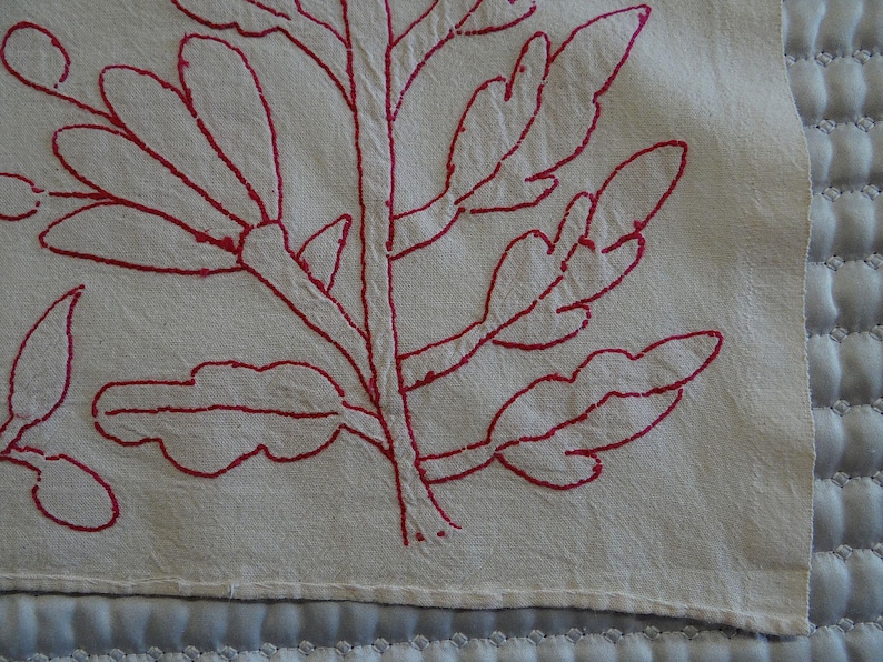 May include: Close-up of a cream fabric with a red embroidered botanical design. The embroidery shows a central stem with branching lines and leaf shapes. The fabric has a textured appearance.