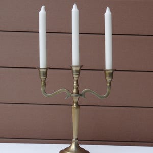Peut inclure: Un chandelier en métal doré à trois bras et une base ronde. Trois bougies blanches sont placées dans les bougeoirs.