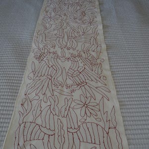K&ouml;nnte beinhalten: Cremefarbener, bestickter Tischl&auml;ufer mit einem sich wiederholenden Muster aus V&ouml;geln, Blumen und stilisierten Tieren in tiefrotem Faden. Das Design ist aufwendig und bedeckt die gesamte L&auml;nge des L&auml;ufers und schafft ein dekoratives Textil.