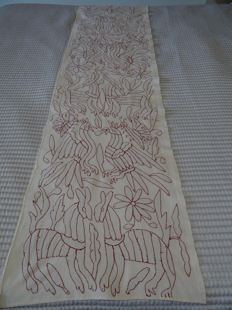 K&ouml;nnte beinhalten: Cremefarbener, bestickter Tischl&auml;ufer mit einem sich wiederholenden Muster aus V&ouml;geln, Blumen und stilisierten Tieren in tiefrotem Faden. Das Design ist aufwendig und bedeckt die gesamte L&auml;nge des L&auml;ufers und schafft ein dekoratives Textil.