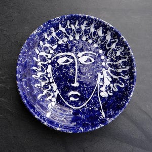 May include: A round, blue ceramic plate with a white face design. The plate has a blue background with white splatter details. The face is drawn with simple white lines, depicting eyes, a nose, and a mouth.