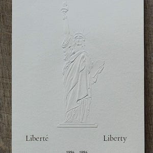 May include: A white embossed card with a depiction of the Statue of Liberty. The statue is shown holding a torch and a tablet. The words "Liberté" and "Liberty" are printed below the statue, with the dates "1886 - 1986".