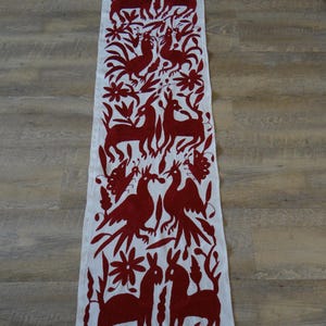 K&ouml;nnte beinhalten: Wei&szlig;er rechteckiger Tischl&auml;ufer mit einem sich wiederholenden Muster aus rot gestickten Tieren, V&ouml;geln und floralen Designs. Der L&auml;ufer ist aus einem weichen Stoff gefertigt und eignet sich f&uuml;r einen Esstisch oder andere Oberfl&auml;chen.