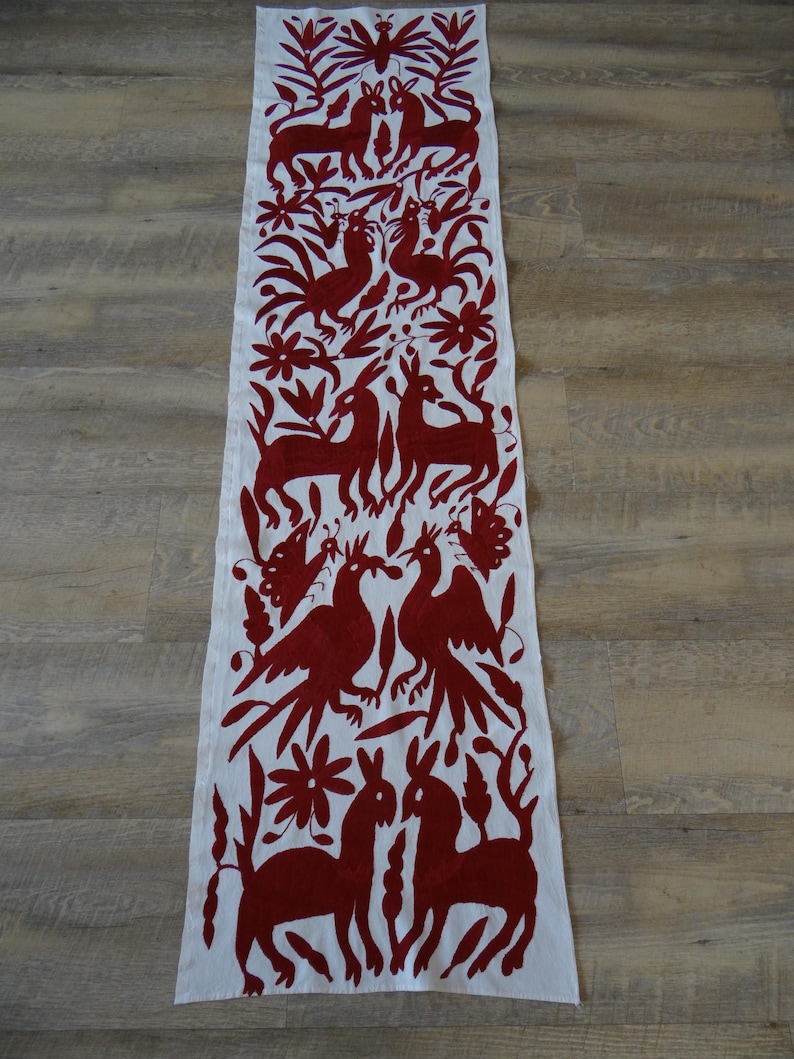 K&ouml;nnte beinhalten: Wei&szlig;er rechteckiger Tischl&auml;ufer mit einem sich wiederholenden Muster aus rot gestickten Tieren, V&ouml;geln und floralen Designs. Der L&auml;ufer ist aus einem weichen Stoff gefertigt und eignet sich f&uuml;r einen Esstisch oder andere Oberfl&auml;chen.