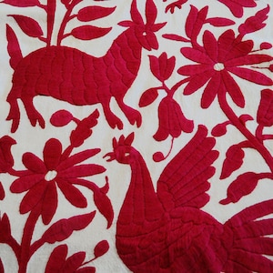May include: White textile with striking red embroidery. The design includes a deer-like creature, a rooster, and floral motifs. The detailed stitching creates a textured pattern. The textile showcases a traditional craft.