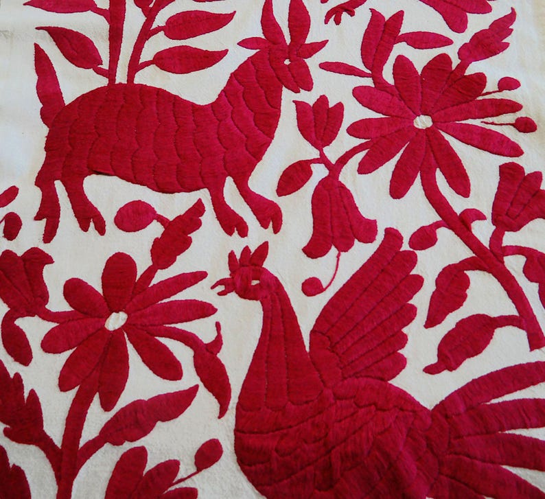 May include: White textile with striking red embroidery. The design includes a deer-like creature, a rooster, and floral motifs. The detailed stitching creates a textured pattern. The textile showcases a traditional craft.