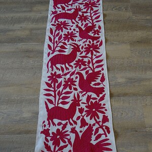 May include: A white table runner with a vibrant magenta embroidered design. The design features stylized animals, flowers, and foliage. The runner is rectangular and appears to be made of a natural fabric.