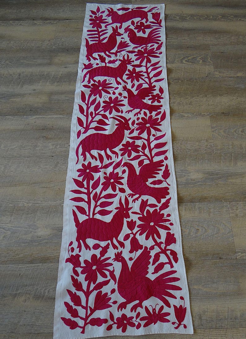May include: A white table runner with a vibrant magenta embroidered design. The design features stylized animals, flowers, and foliage. The runner is rectangular and appears to be made of a natural fabric.