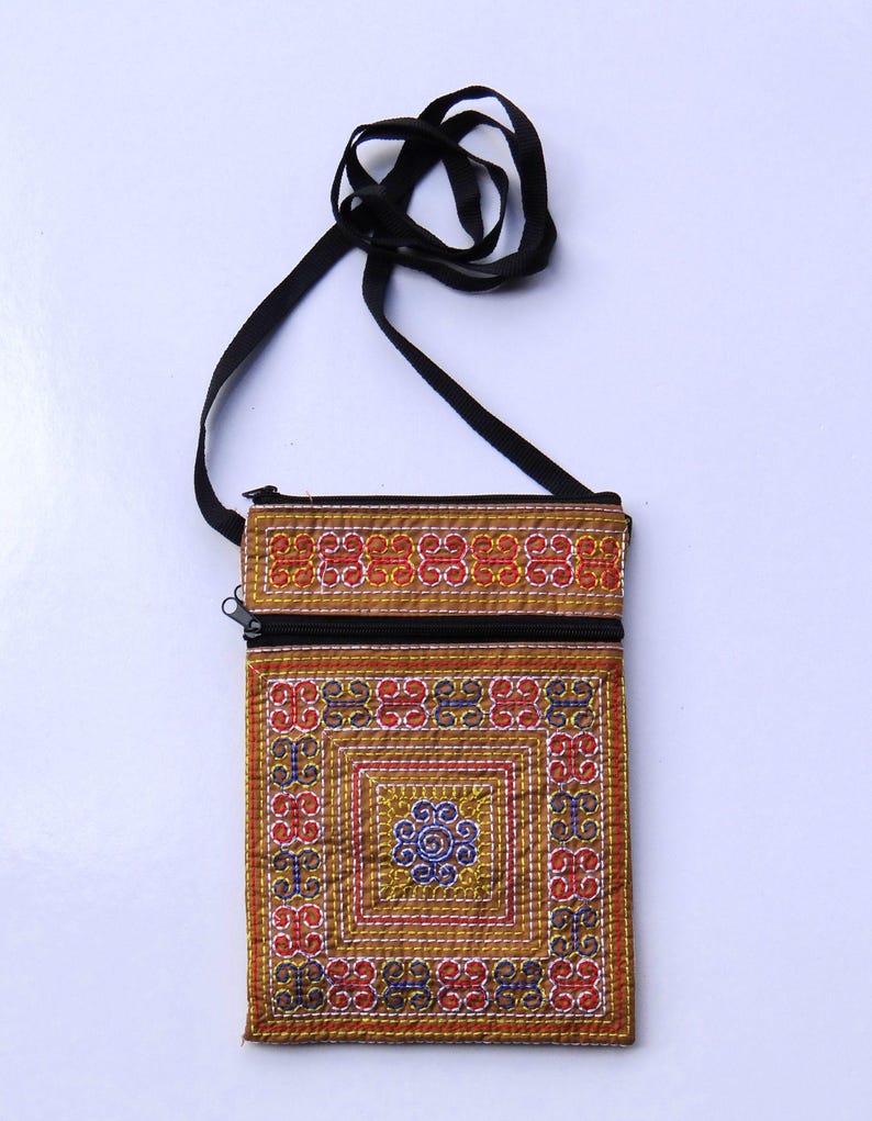 Puede incluir: Un peque&ntilde;o bolso bandolera rectangular bordado con una correa negra. El bolso presenta un colorido patr&oacute;n geom&eacute;trico en rojo, azul y dorado. Una cremallera negra recorre la parte superior. Es probable que el bolso est&eacute; hecho a mano.