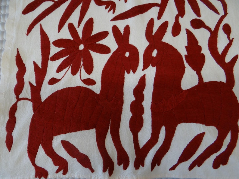 K&ouml;nnte beinhalten: Wei&szlig;er Stoff mit einem roten Stickmuster, das zwei stilisierte Tiere zeigt, die sich gegen&uuml;berstehen, umgeben von Blumen- und Blattmotiven. Die Stickerei ist detailliert und das Design ist symmetrisch.