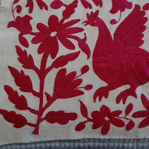 May include: Embroidered textile with a vibrant fuchsia design on an off-white background. The design features a stylized bird, flowers, and foliage. The embroidery is detailed and creates a textured appearance, showcasing a folk art aesthetic.