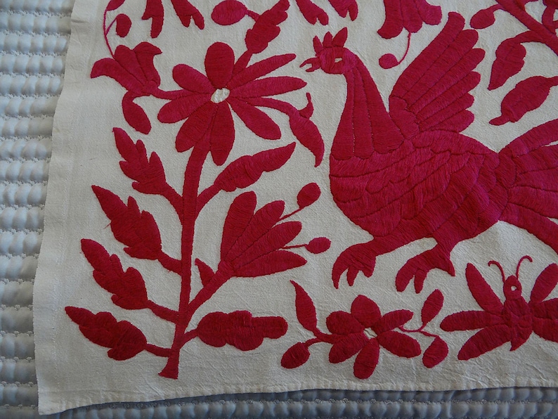 May include: Embroidered textile with a vibrant fuchsia design on an off-white background. The design features a stylized bird, flowers, and foliage. The embroidery is detailed and creates a textured appearance, showcasing a folk art aesthetic.