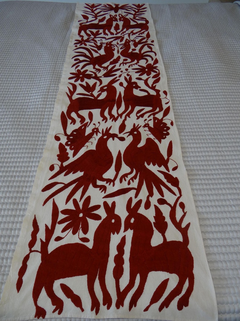 K&ouml;nnte beinhalten: Cremefarbener Tischl&auml;ufer mit einem sich wiederholenden Muster aus stilisierten Tieren und floralen Elementen in einem tiefroten Farbton. Das Design umfasst Hirsche, V&ouml;gel und Blumen und erzeugt eine Volkskunst-&Auml;sthetik. Der L&auml;ufer ist wahrscheinlich aus Stoff.