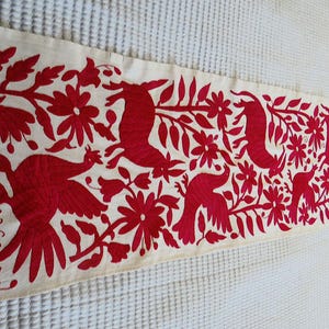 May include: Cream-coloured rectangular table runner with a vibrant red embroidered design. The design features stylised birds, flowers, and foliage. The runner is likely made of a natural fabric, adding a touch of rustic charm to any table setting.
