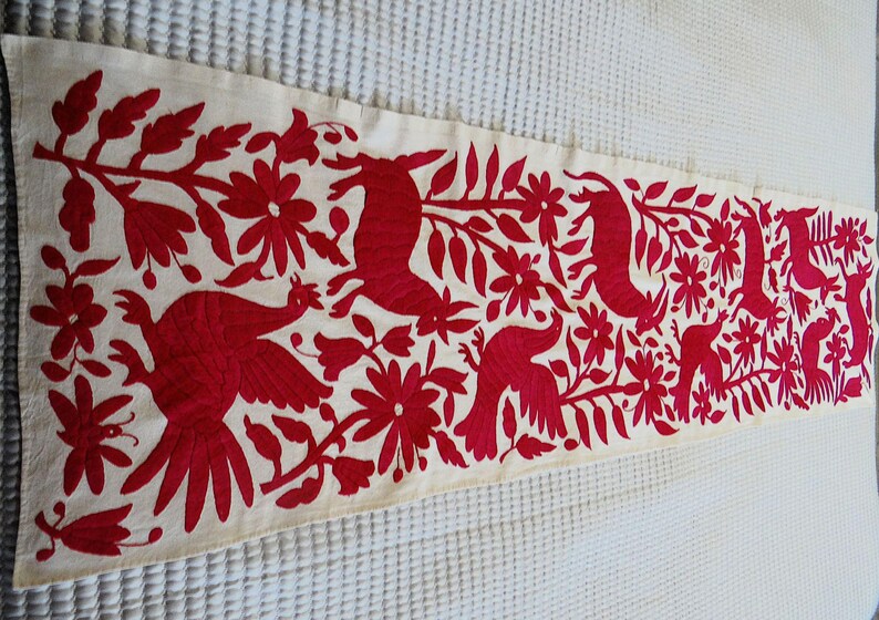 May include: Cream-coloured rectangular table runner with a vibrant red embroidered design. The design features stylised birds, flowers, and foliage. The runner is likely made of a natural fabric, adding a touch of rustic charm to any table setting.