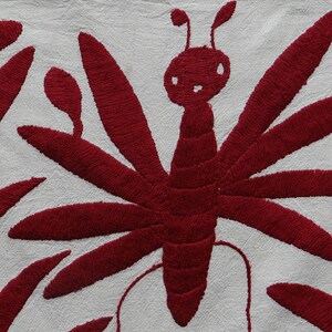 K&ouml;nnte beinhalten: Nahaufnahme eines wei&szlig;en Stoffes mit einem detaillierten Stickdesign. Das Design zeigt einen gro&szlig;en, stilisierten roten Schmetterling mit ausgebreiteten Fl&uuml;geln und eine Reihe roter, blattartiger Formen. Die Stickerei hat ein strukturiertes Aussehen.