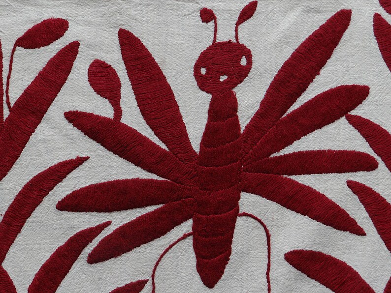 K&ouml;nnte beinhalten: Nahaufnahme eines wei&szlig;en Stoffes mit einem detaillierten Stickdesign. Das Design zeigt einen gro&szlig;en, stilisierten roten Schmetterling mit ausgebreiteten Fl&uuml;geln und eine Reihe roter, blattartiger Formen. Die Stickerei hat ein strukturiertes Aussehen.