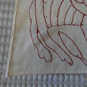 K&ouml;nnte beinhalten: Nahaufnahme eines wei&szlig;en Textils mit einem roten, abstrakten Design. Das Design scheint handgen&auml;ht oder gestickt zu sein. Das Textil liegt auf einer wei&szlig;en, gesteppten Oberfl&auml;che.