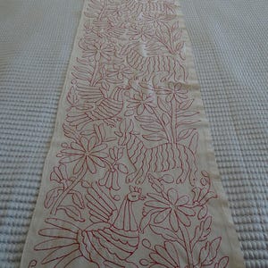 May include: Cream-coloured table runner with red embroidered floral and animal designs. The runner features detailed stitching of birds, flowers, and other botanical elements, creating a decorative textile for home decor.