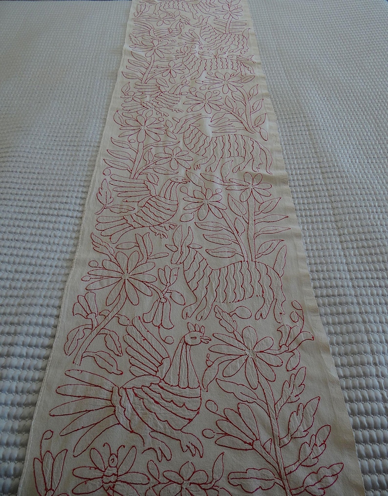 May include: Cream-coloured table runner with red embroidered floral and animal designs. The runner features detailed stitching of birds, flowers, and other botanical elements, creating a decorative textile for home decor.