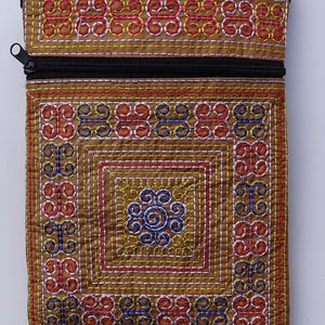 Puede incluir: Una bolsa rectangular de tela marr&oacute;n con cierre de cremallera negro. La bolsa est&aacute; decorada con intrincados patrones bordados en hilo rojo, azul, amarillo y blanco, creando un dise&ntilde;o geom&eacute;trico. El dise&ntilde;o incluye un motivo cuadrado central.