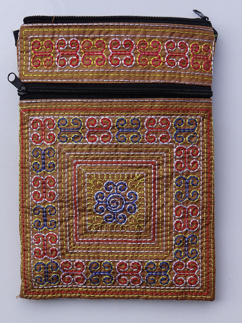 Puede incluir: Una bolsa rectangular de tela marr&oacute;n con cierre de cremallera negro. La bolsa est&aacute; decorada con intrincados patrones bordados en hilo rojo, azul, amarillo y blanco, creando un dise&ntilde;o geom&eacute;trico. El dise&ntilde;o incluye un motivo cuadrado central.