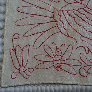 May include: Close-up of a cream textile with red embroidered floral and insect designs. The embroidery features a butterfly, flowers, and other stylized floral elements. The textile is likely a decorative piece.