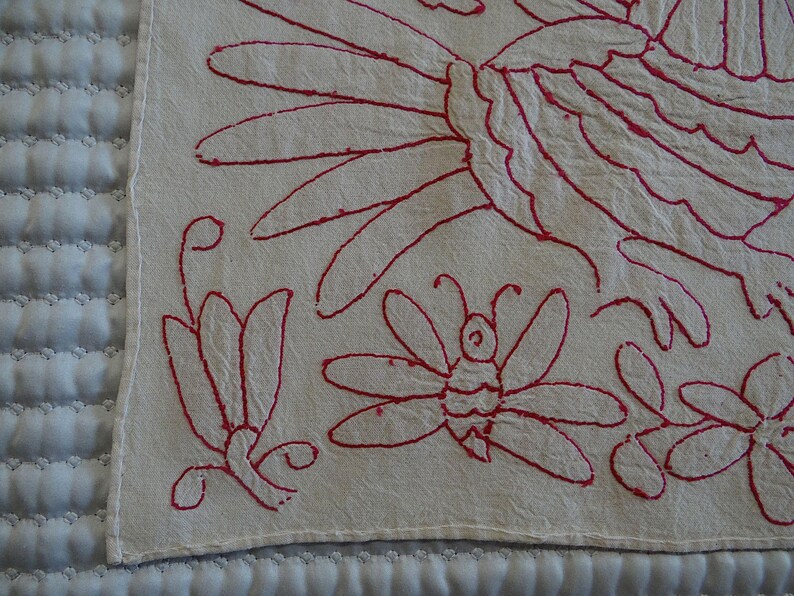May include: Close-up of a cream textile with red embroidered floral and insect designs. The embroidery features a butterfly, flowers, and other stylized floral elements. The textile is likely a decorative piece.