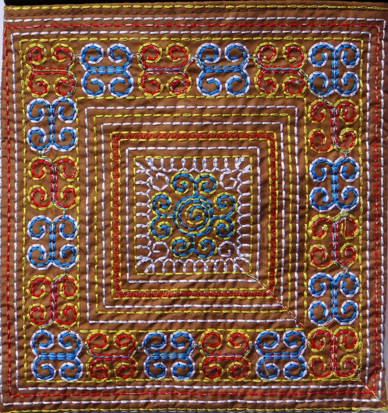 Puede incluir: Textil cuadrado con intrincado bordado en marr&oacute;n, rojo, amarillo, blanco y azul. El dise&ntilde;o presenta patrones geom&eacute;tricos repetidos y un motivo floral central. La costura es detallada y crea una superficie texturizada.
