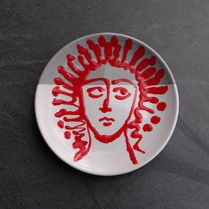May include: A white ceramic plate with a red and gray abstract face design. The face is outlined in red, with a gray background on the top half of the plate. The plate is round and has a simple, modern aesthetic.
