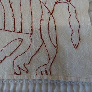 K&ouml;nnte beinhalten: Nahaufnahme eines cremefarbenen Textils mit einem roten, handgezeichneten Design. Das Design scheint eine abstrakte Darstellung einer Figur oder Figuren zu sein. Der Stoff hat eine leicht zerknitterte Textur und die Kanten sind sauber verarbeitet.
