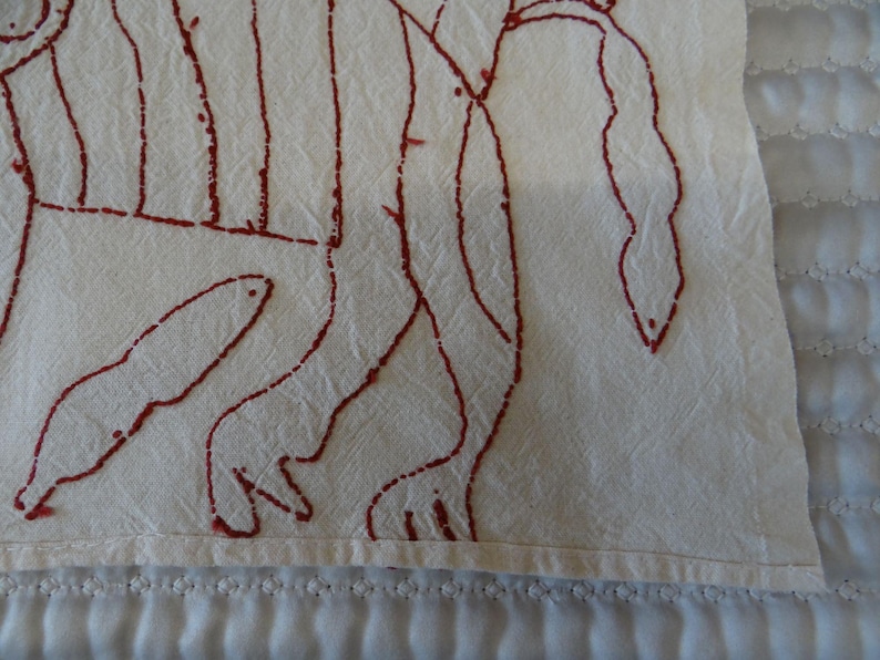 K&ouml;nnte beinhalten: Nahaufnahme eines cremefarbenen Textils mit einem roten, handgezeichneten Design. Das Design scheint eine abstrakte Darstellung einer Figur oder Figuren zu sein. Der Stoff hat eine leicht zerknitterte Textur und die Kanten sind sauber verarbeitet.
