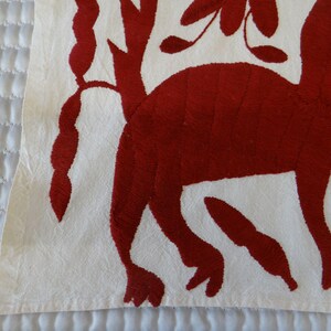 K&ouml;nnte beinhalten: Nahaufnahme eines wei&szlig;en Stoffes mit rotem Stickmuster. Das Design zeigt eine stilisierte Tierfigur und florale Elemente. Der Stoff hat ein strukturiertes Aussehen, wobei die rote Stickerei vor dem wei&szlig;en Hintergrund hervorsticht.