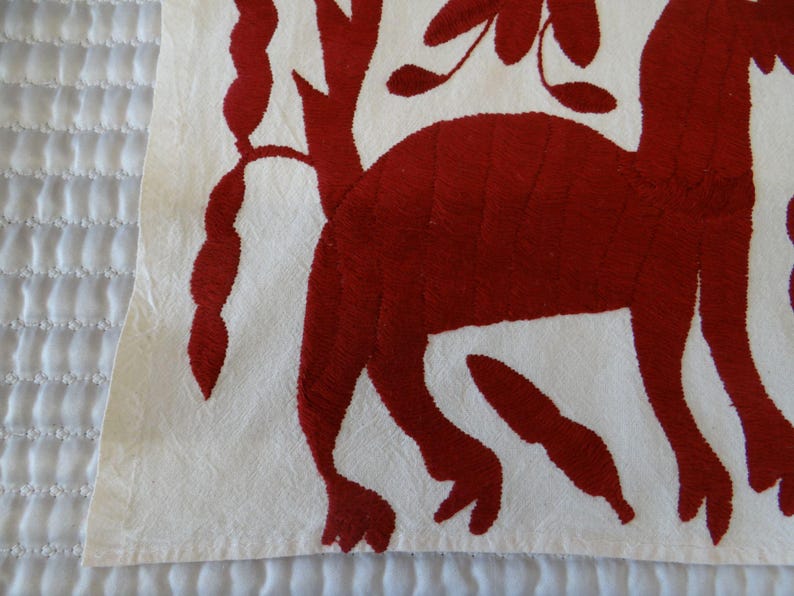K&ouml;nnte beinhalten: Nahaufnahme eines wei&szlig;en Stoffes mit rotem Stickmuster. Das Design zeigt eine stilisierte Tierfigur und florale Elemente. Der Stoff hat ein strukturiertes Aussehen, wobei die rote Stickerei vor dem wei&szlig;en Hintergrund hervorsticht.