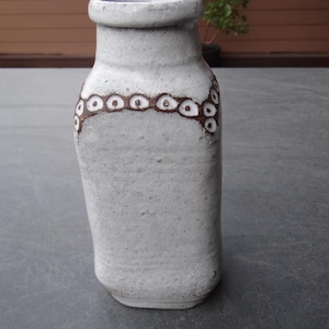 Könnte beinhalten: Eine handgefertigte, cremefarbene Keramikvase mit strukturierter Oberfläche. Die Vase hat ein dekoratives Band aus braunen, kreisförmigen Akzenten um den Hals. Die Vase hat einen rechteckigen Körper und einen abgerundeten Boden. Ein Unikat für die Inneneinrichtung.