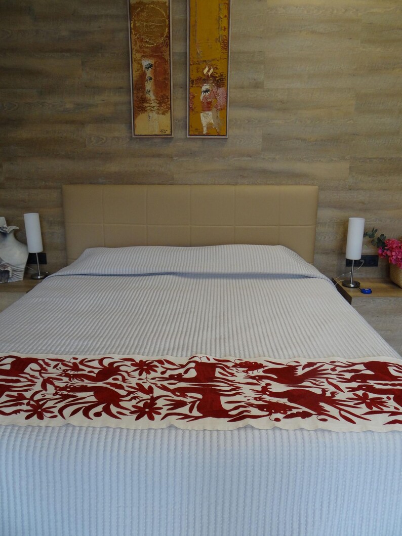 K&ouml;nnte beinhalten: Ein Bett mit einer hellblau-wei&szlig; gestreiften Bettdecke und einem dekorativen rot-wei&szlig;en L&auml;ufer. Das Kopfteil ist beige, und zwei gerahmte Gem&auml;lde h&auml;ngen &uuml;ber dem Bett. Zwei wei&szlig;e Lampen stehen auf den Nachttischen.