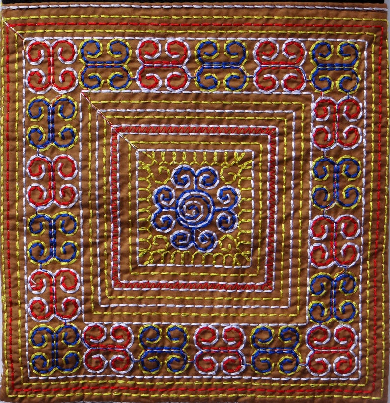 Puede incluir: Textil cuadrado con intrincados bordados en tonos marr&oacute;n, rojo, amarillo, azul y blanco. El dise&ntilde;o presenta un motivo floral central y patrones geom&eacute;tricos repetidos en los bordes. La costura es detallada y precisa.