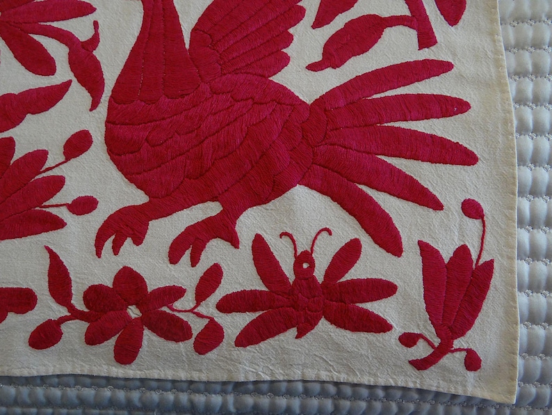 May include: Embroidered textile with a red rooster, flowers, and a butterfly on an off-white background. The detailed embroidery creates a textured surface, showcasing a folk art style. The design is likely part of a larger piece.