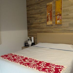 May include: A bedroom with a bed covered in a white and blue striped duvet. A decorative red and cream-coloured runner with floral and bird designs is placed at the foot of the bed. Two abstract paintings hang on the wall above the bed.