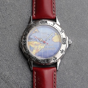 May include: A silver wristwatch with a red leather band. The watch face features a world map design with the text "Sur les traces de l'Aéropostale" and the years 1930 and 1998. The watch has a silver bezel with numbers.