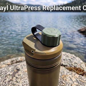May include: A tan Grayl UltraPress replacement cap featuring a black loop handle and a small olive green screw-on lid with a triangular logo. The cap has a textured top surface and a small metal eyelet on the side, designed for a water bottle.